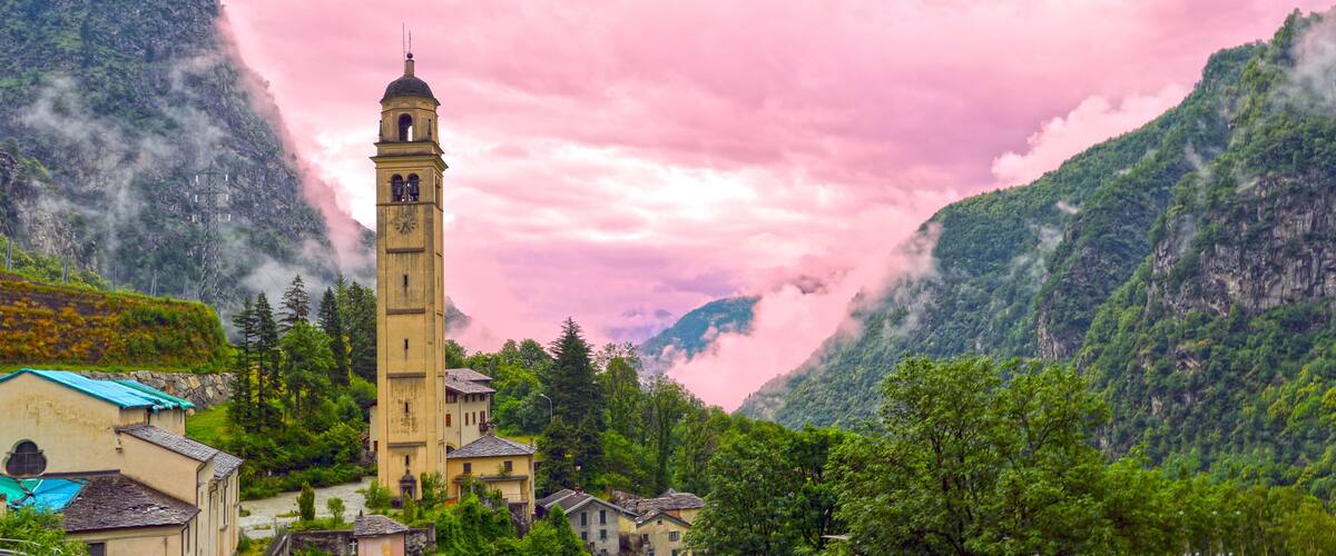 A church with a bell tower surrounded by lush green mountains and a pink sky.