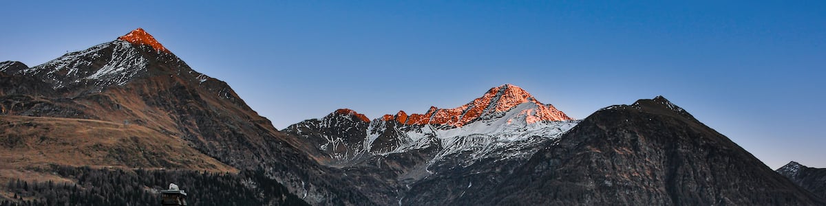 Ein Bergdorf mit Steinhäusern und einem markanten Gipfel mit orangen Schneeflecken.
