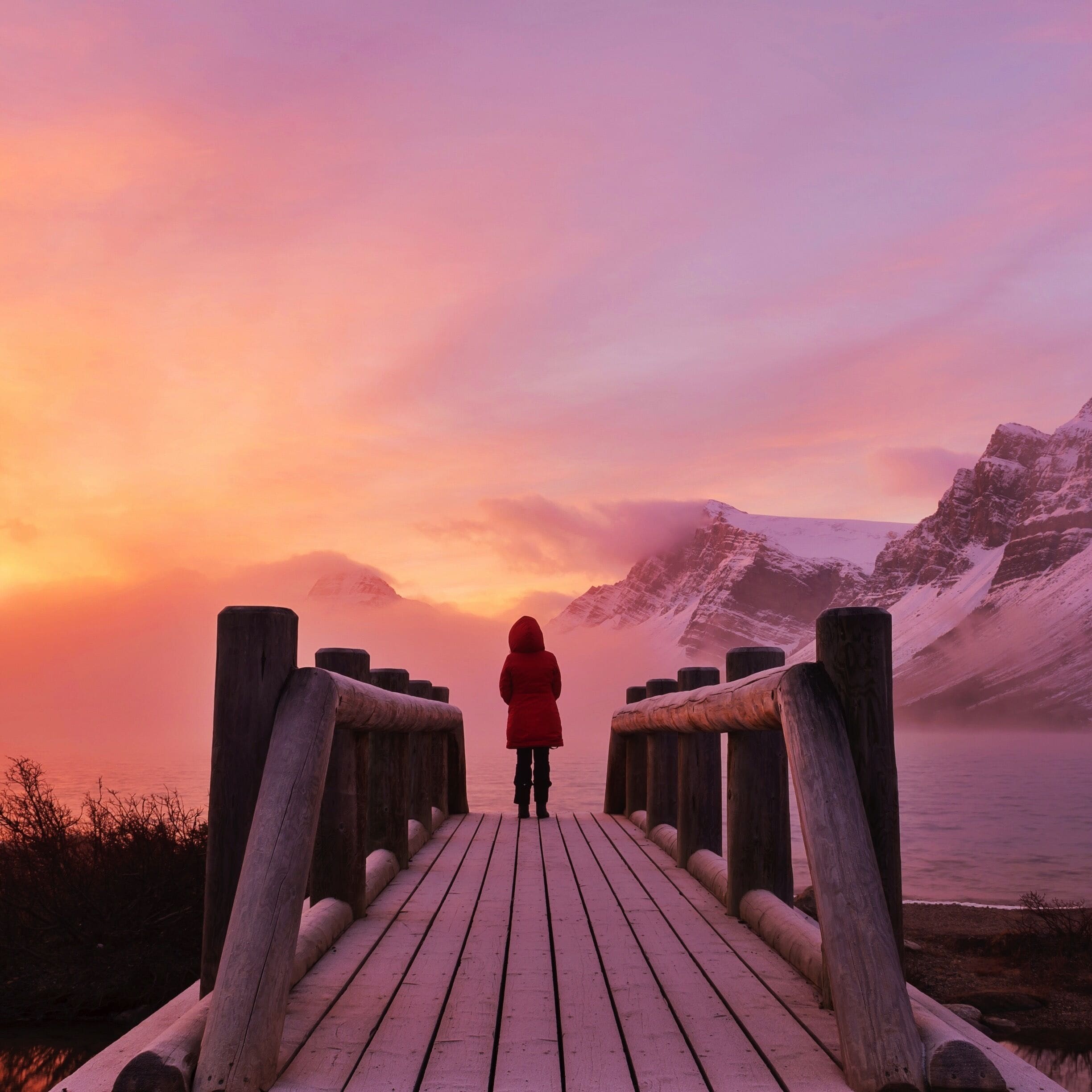 A wooden boardwalk leading to a snowy mountain landscape at sunset.