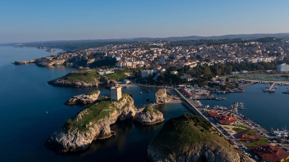 A coastal cityscape with a prominent castle on a rocky outcrop, a marina with boats, and a densely built urban area in the background.