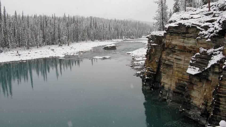 A snow-covered river with a rocky cliff and dense forest.
