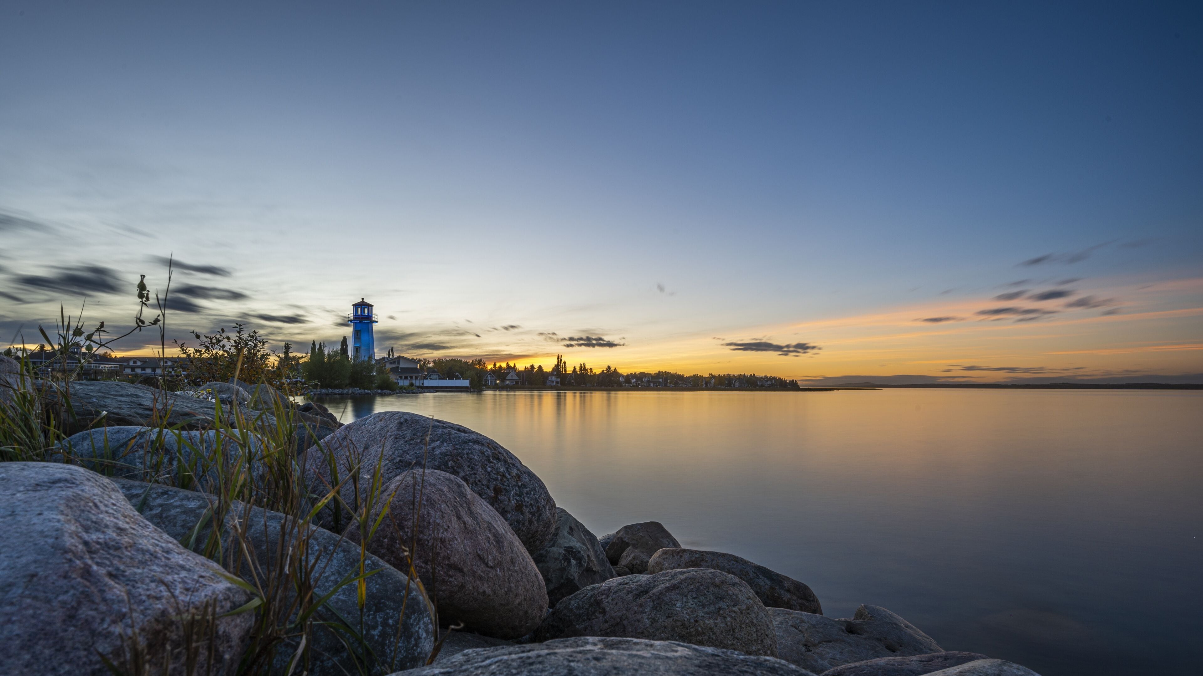 A calm lake at sunset with a lighthouse and rocky shoreline.