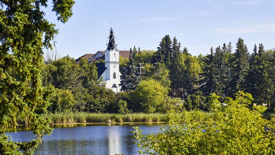 A white church with a cross on top, surrounded by trees and a lake.