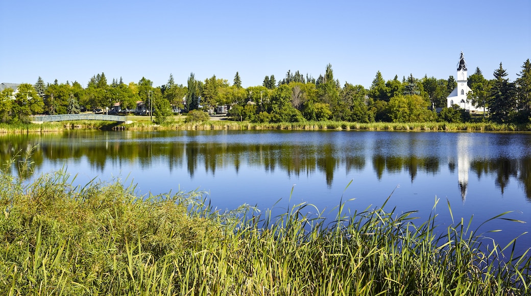 Tranquil Mirror Lake in the Camrose community with a footbridge from a park and the white Ukrainian Catholic church building reflected in the water; Camrose, Alberta, Canada
