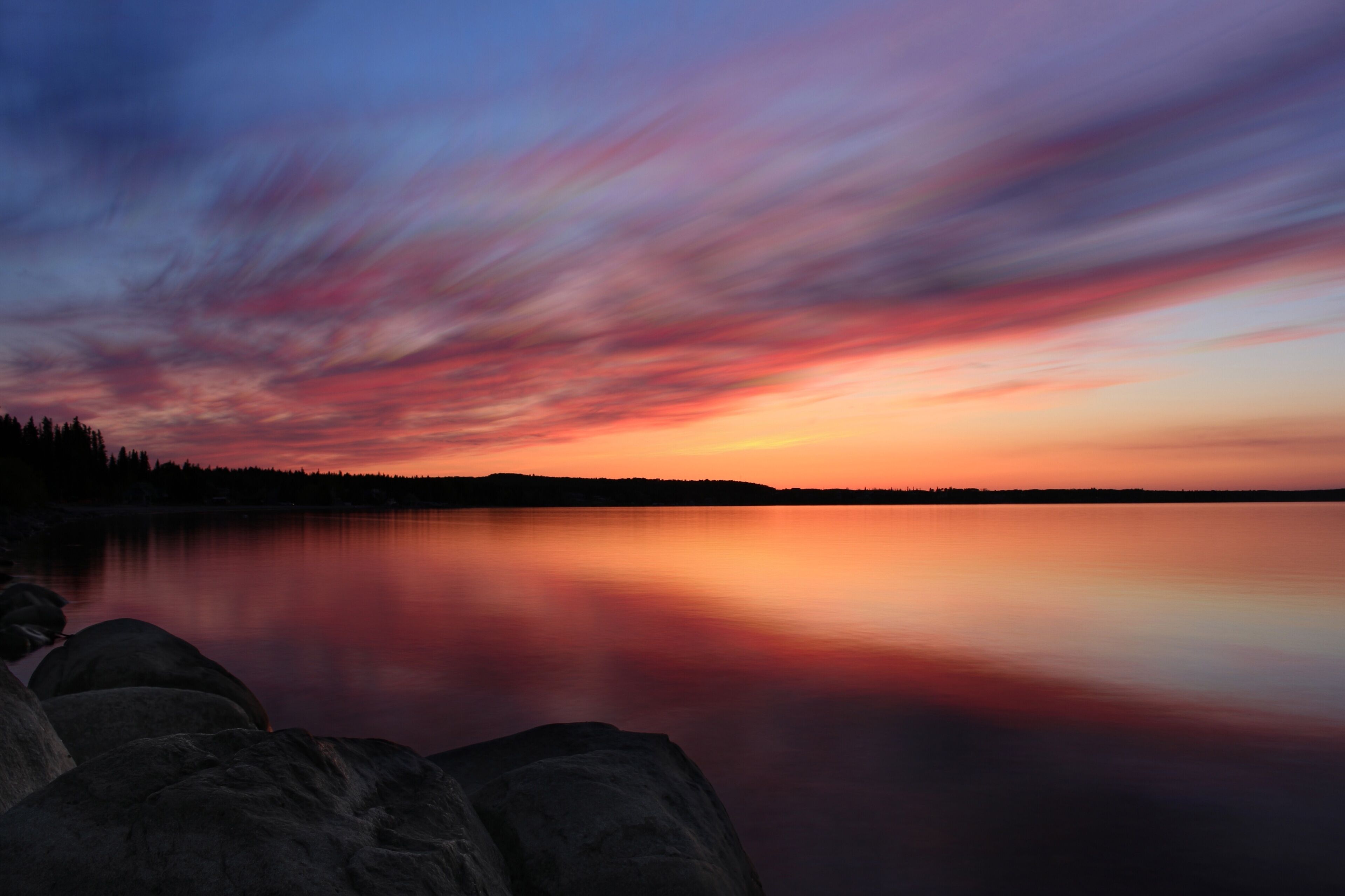 A sunset over a calm lake with a silhouette of trees on the horizon.