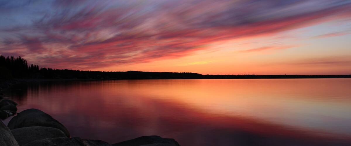 A sunset over a calm lake with a silhouette of trees on the horizon.