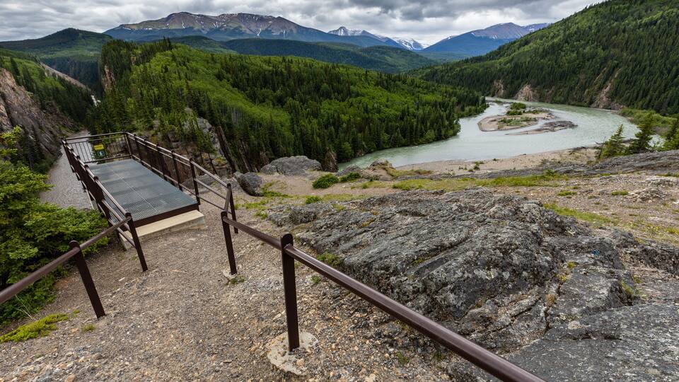 A wooden viewing platform overlooking a river, surrounded by dense forest and mountains.