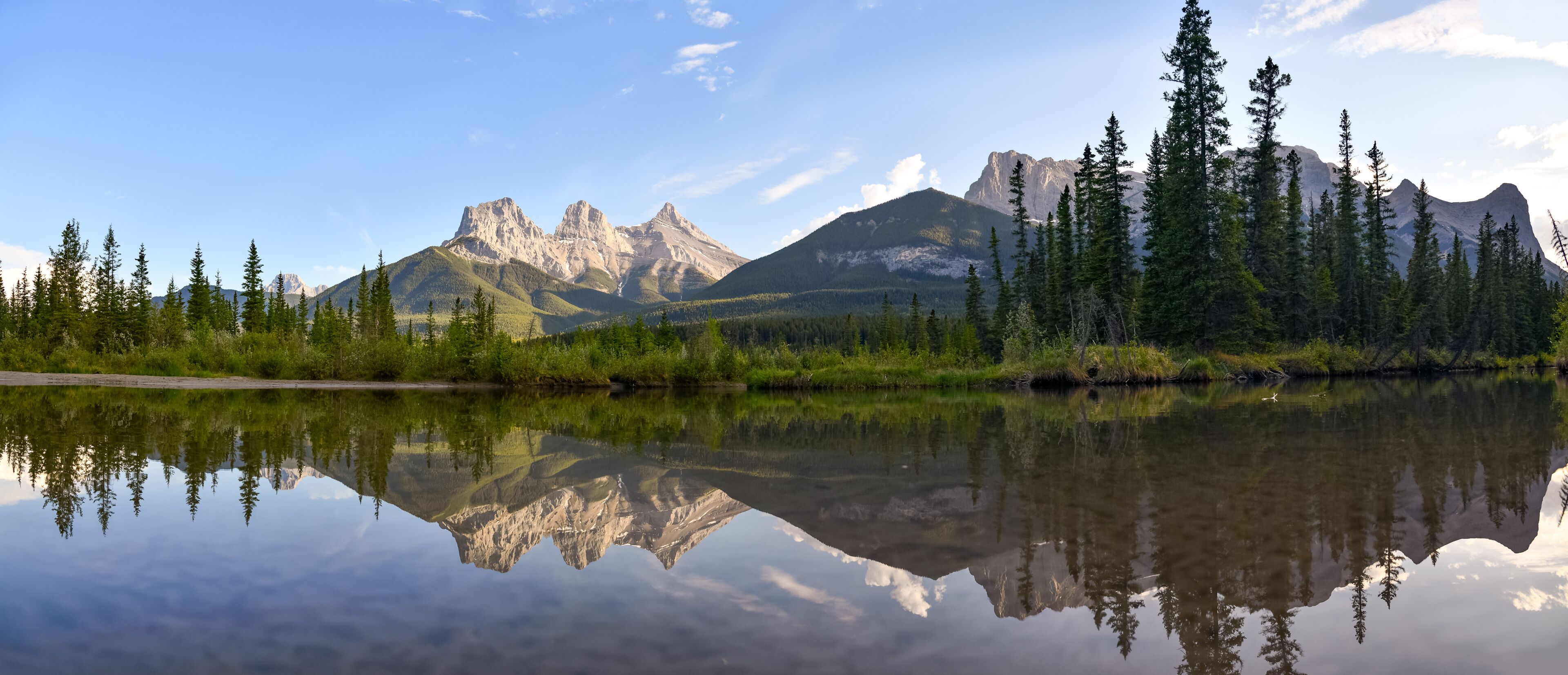 Three Sisters in Canmore seen at golden hour, sunset on blue sky day, afternoon with calm, peaceful reflection in water below famous, tourist, tourism mountains, area summer.	