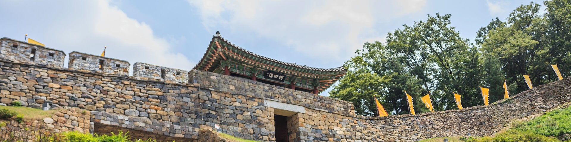 Uma fortaleza coreana tradicional com muros de pedra e telhado de telhas, rodeada por vegetação exuberante e árvores.