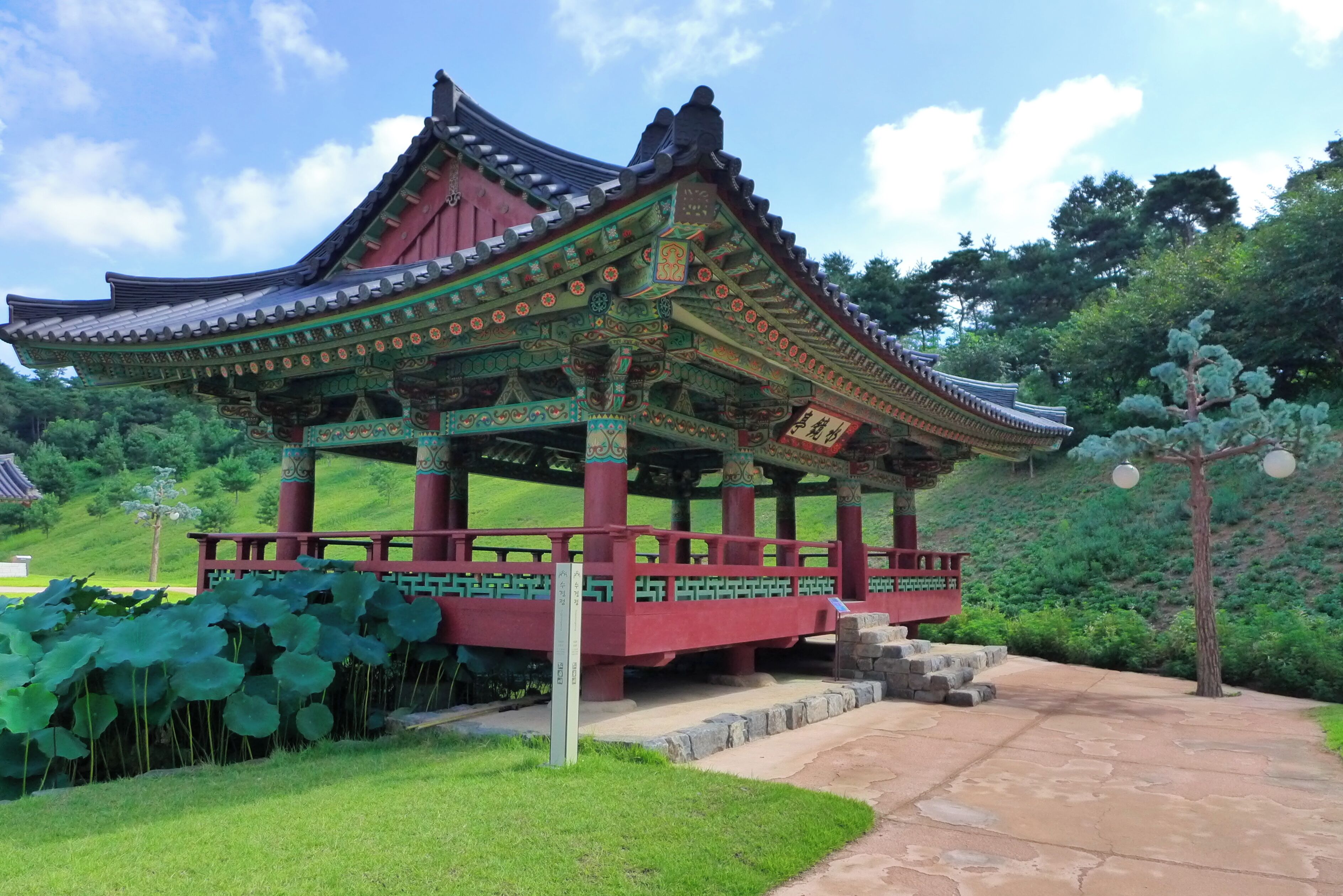 Um pavilhão coreano tradicional com telhado e colunas ricamente decorados, rodeado por vegetação exuberante e um caminho de pedra.