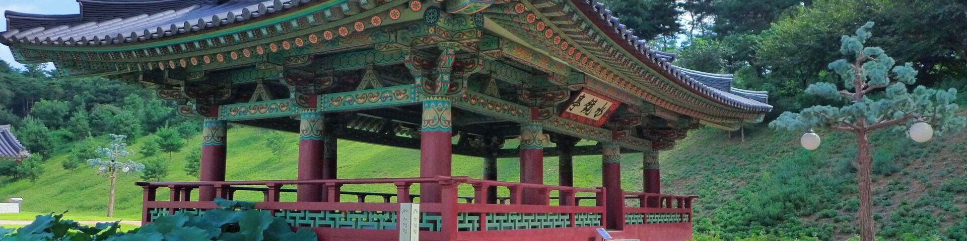 Um pavilhão coreano tradicional com telhado e colunas ricamente decorados, rodeado por vegetação exuberante e um caminho de pedra.