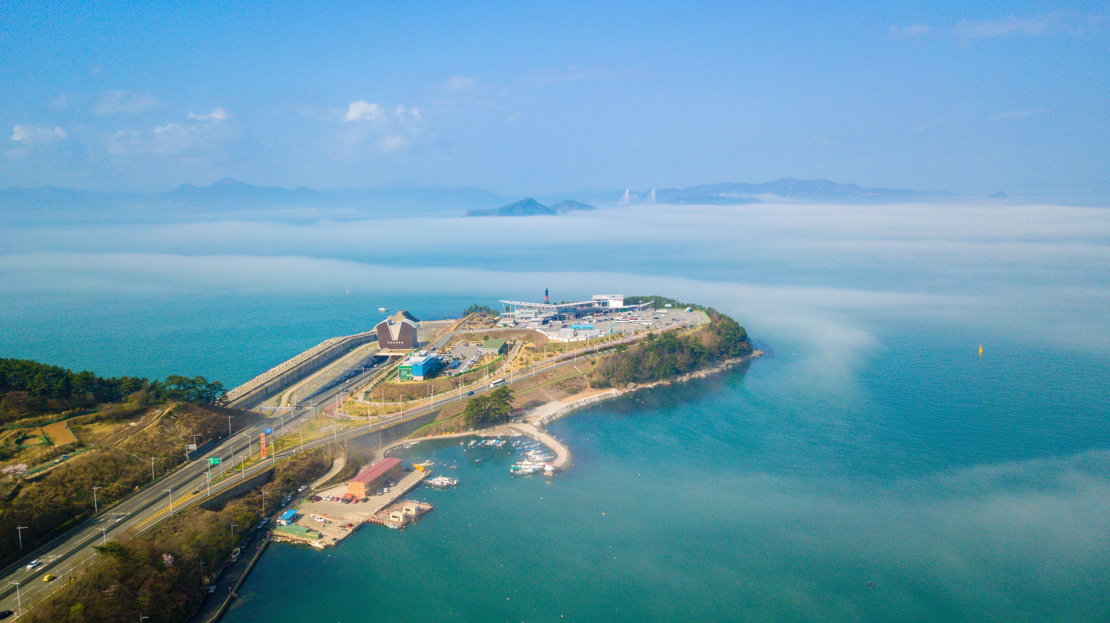An aerial view of a coastal area with a bridge, a lighthouse, and a peninsula with buildings and roads.