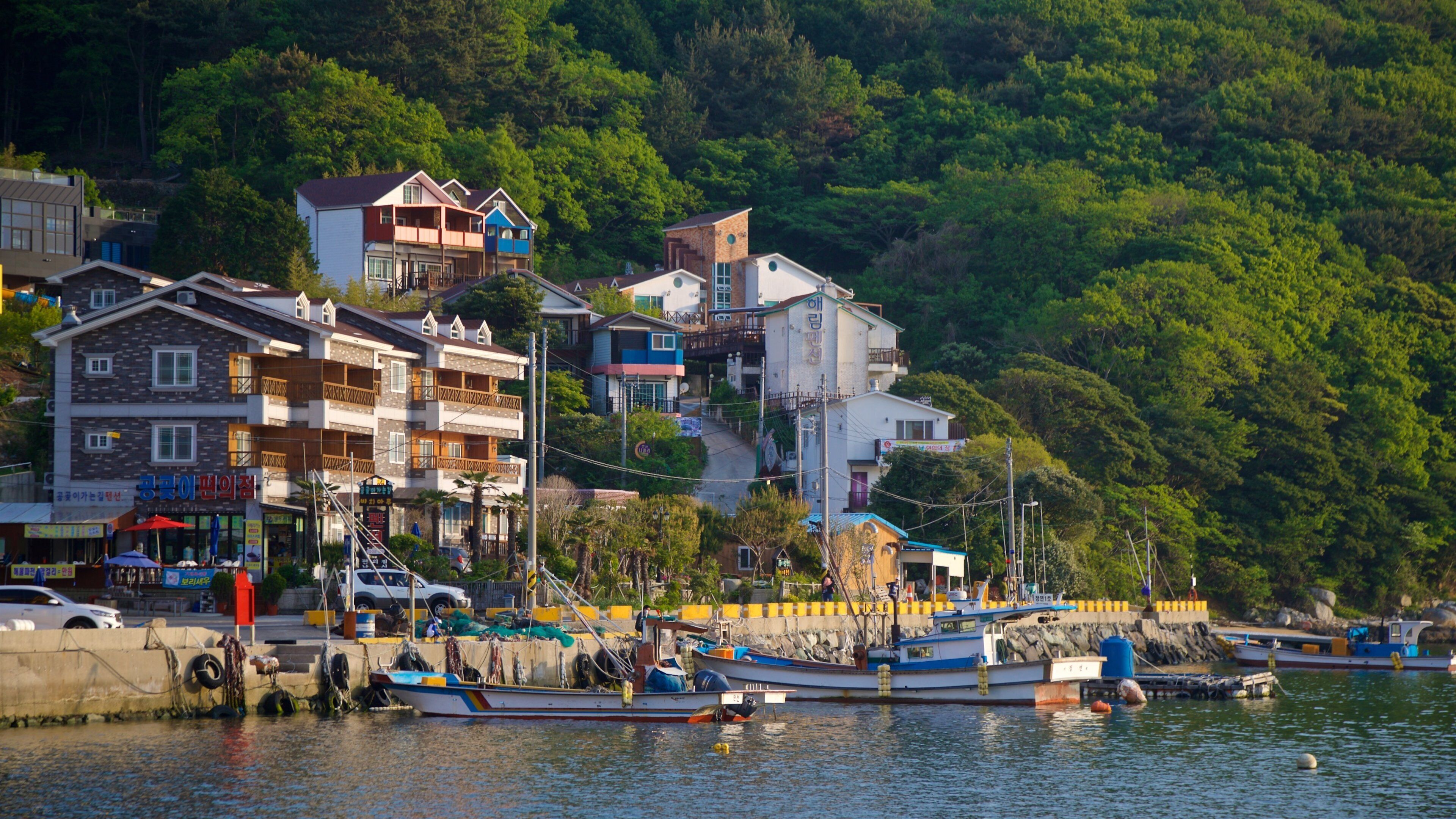 A coastal village with multi-story buildings, a dock with boats, and lush green hills in the background.