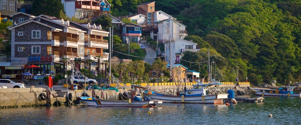 A coastal village with multi-story buildings, a dock with boats, and lush green hills in the background.