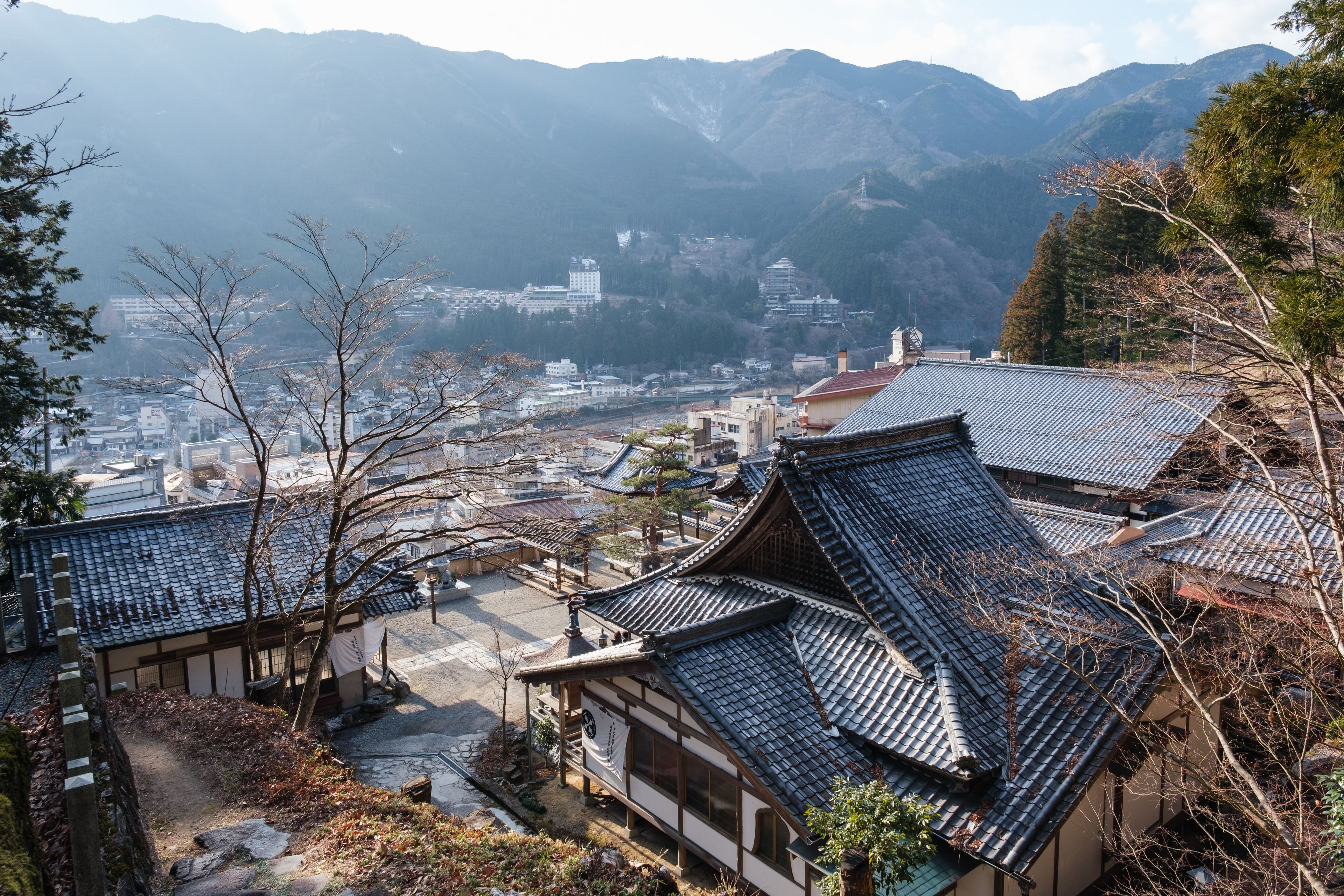 Traditional Japanese buildings with tiled roofs, a mountainous backdrop, and a distant view of a cityscape.