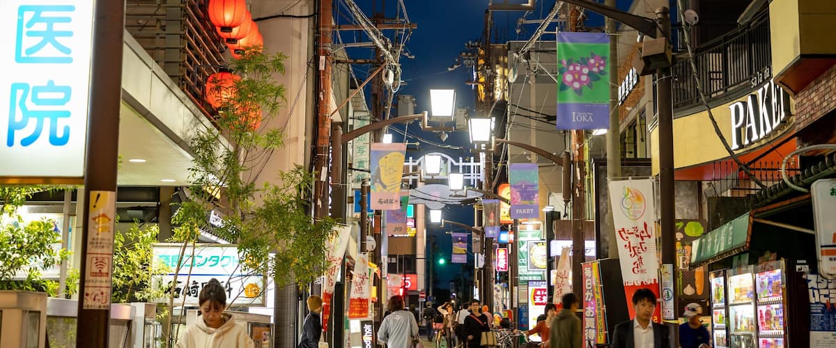 A busy urban street at night with illuminated signs, lanterns, and various shops.