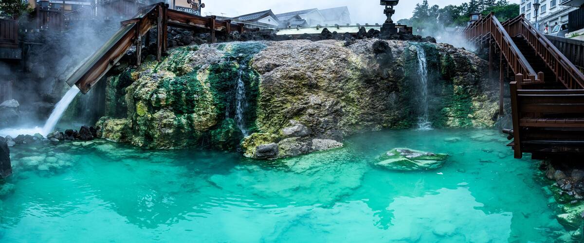 A traditional Japanese onsen with clear turquoise water, wooden walkways, and a wooden bridge.