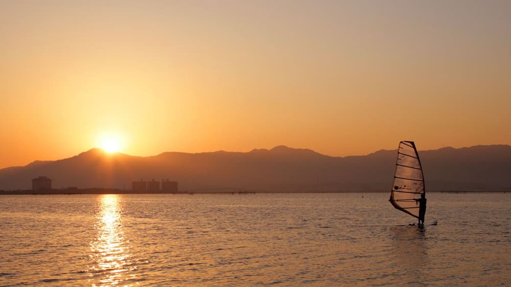 Un coucher de soleil sur une étendue d’eau calme, avec en arrière-plan la silhouette de montagnes et une personne pratiquant la planche à voile.