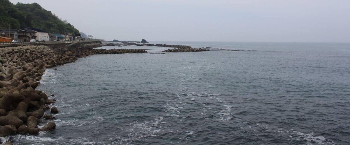 A rocky shoreline with waves crashing against it, a bridge, and buildings in the background.