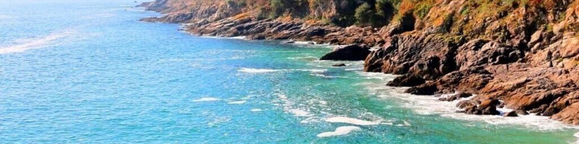 A coastal landscape with clear blue waters, rocky cliffs, and dense forest.