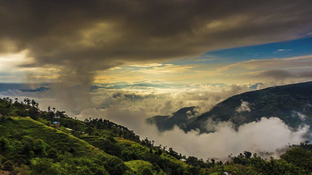 A mountainous landscape with lush green hills, a valley filled with clouds, and a dramatic sky with dark clouds and sunlight breaking through.