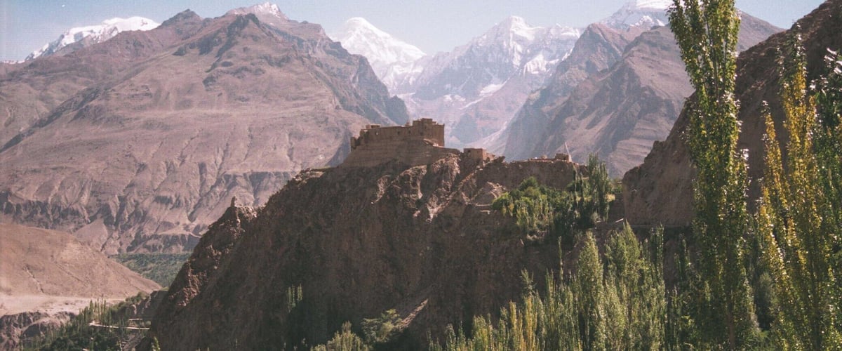 A mountainous landscape with snow-capped peaks, a rocky outcrop with ruins, and dense green vegetation.