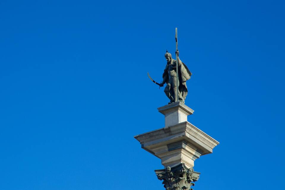 Una estatua de una figura sosteniendo una espada, montada sobre un pedestal con intrincados grabados, contra un cielo azul despejado.