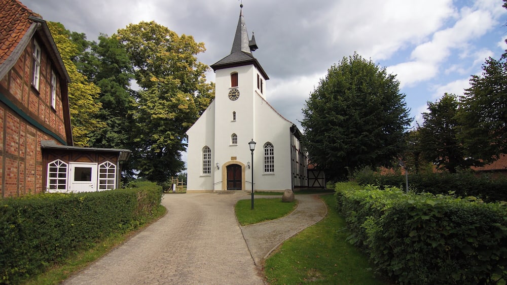 Une église dotée d’un clocher imposant, un bâtiment en briques sur la gauche et un chemin de gravier menant à l’entrée.