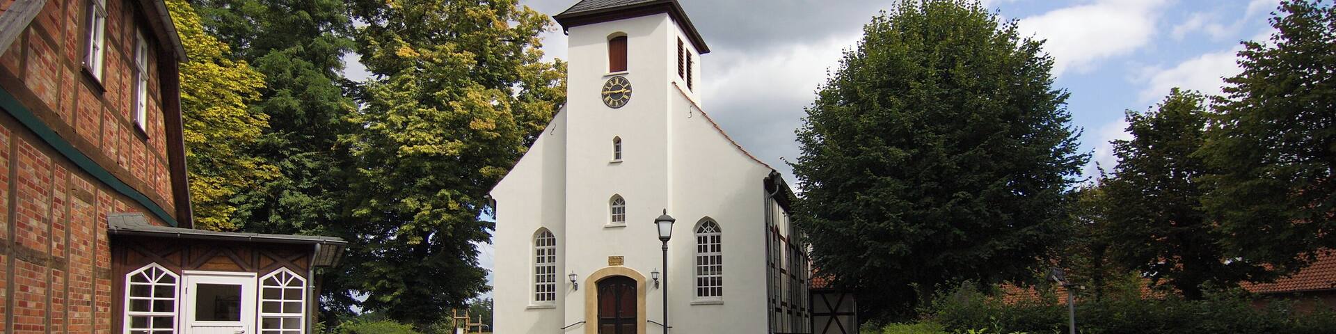 A church with a tall steeple, a brick building to the left, and a gravel path leading to the entrance.