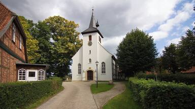 Uma igreja com uma torre alta, um edifício de tijolos à esquerda e um caminho de cascalho que leva à entrada.