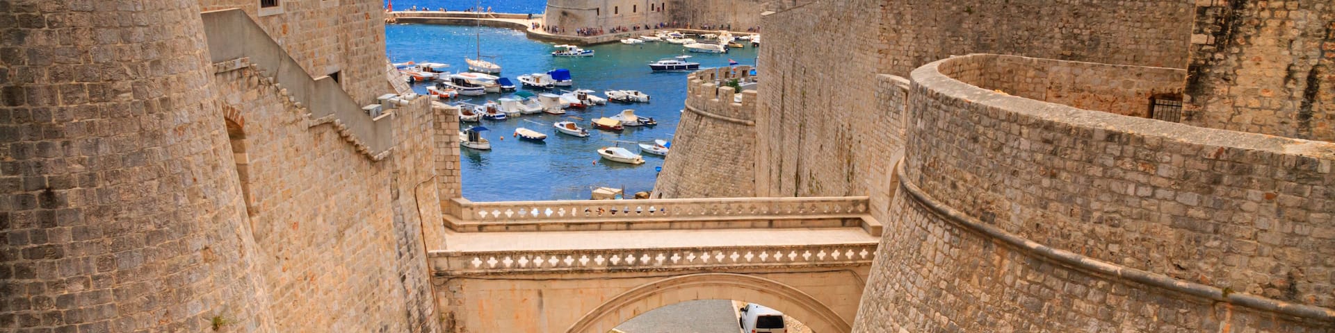 A stone bridge over a moat, surrounded by historic stone walls and a marina with boats.