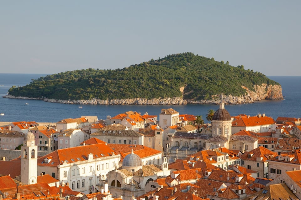 A coastal town with red-roofed buildings, a prominent church with a dome, and a small island with dense greenery in the background.