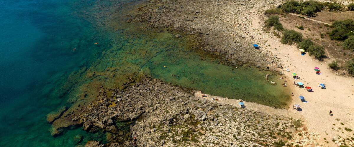 A rocky coastline with clear blue waters, a sandy beach with umbrellas, and a small boat in the distance.