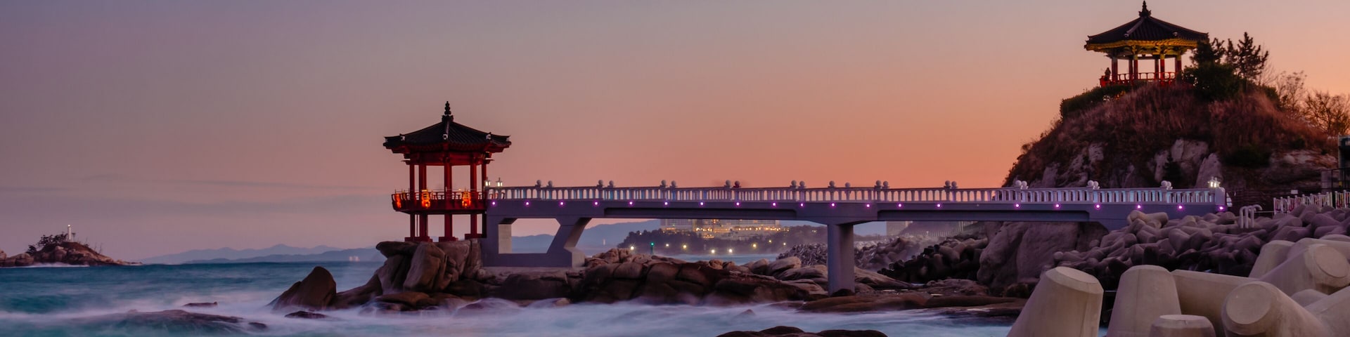 Uma paisagem costeira com um gazebo de madeira, uma ponte de pedra e uma linha costeira rochosa.