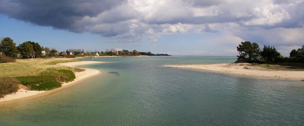 A coastal landscape with a sandy beach, calm waters, and a distant shoreline with trees and houses.