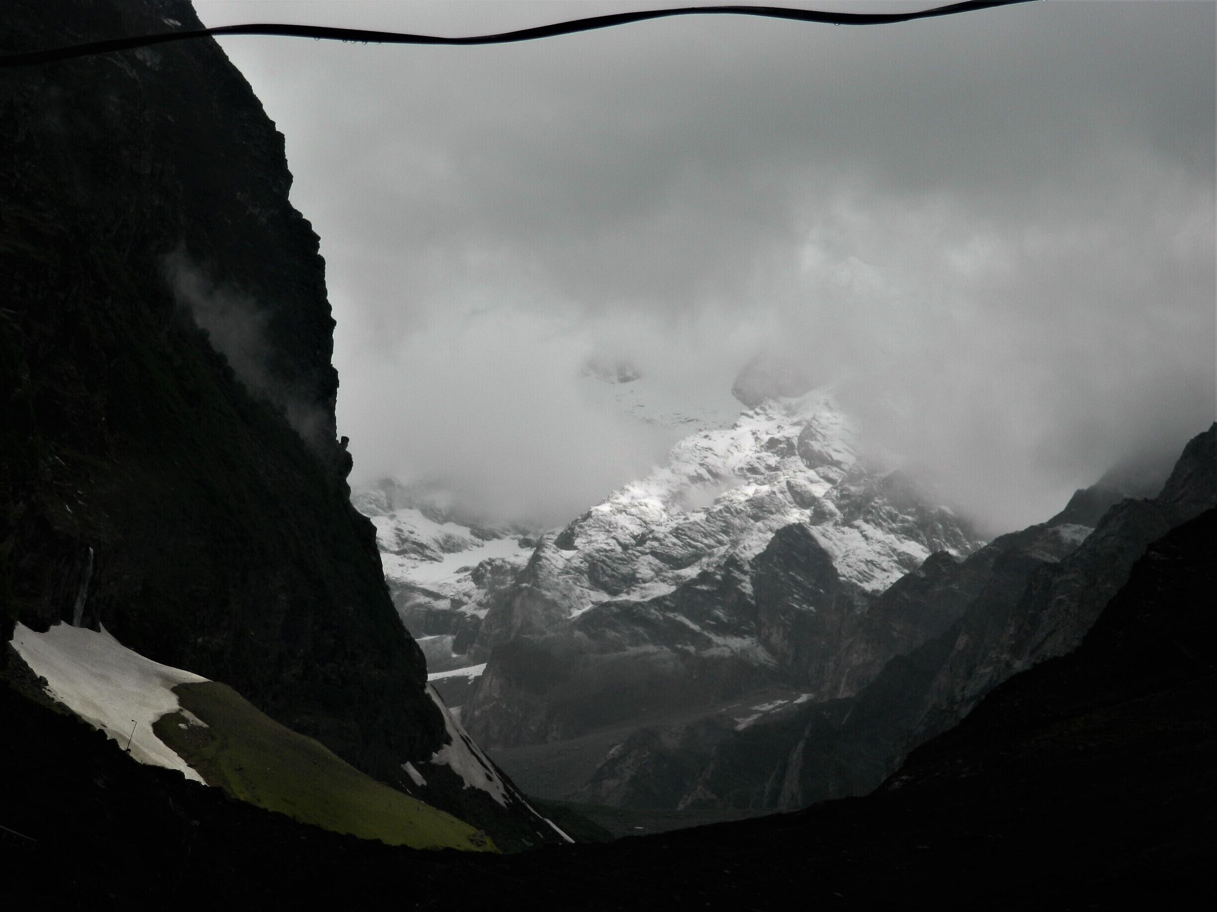 A snow-covered mountain peak partially obscured by clouds, with a rocky foreground and a wireframe in the upper left corner.