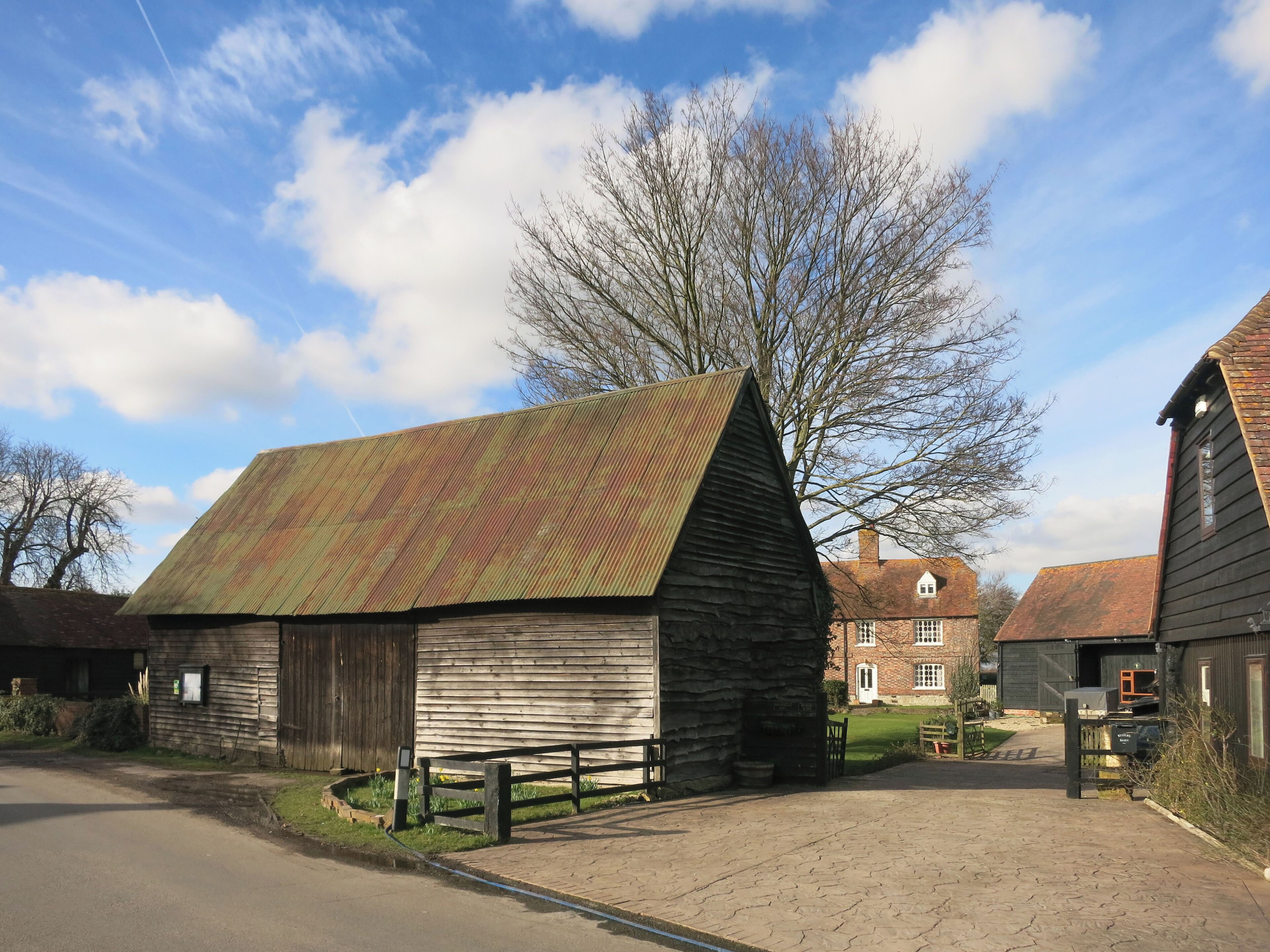 A rustic barn with a sloped roof, surrounded by a paved driveway and a large tree.