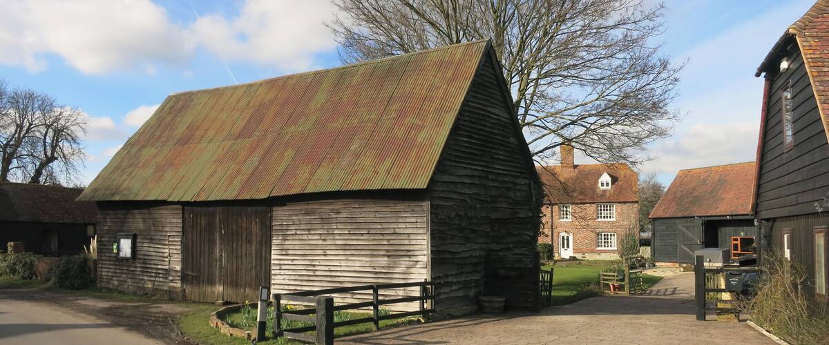 A rustic barn with a sloped roof, surrounded by a paved driveway and a large tree.