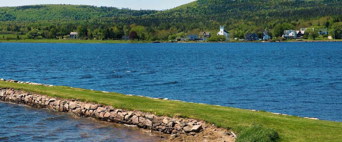 A calm lake with a stone wall and grassy shore. In the background, there are houses and a church on a hill.