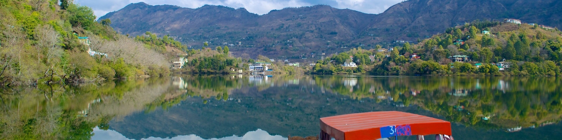 A calm lake with a boat docked on the right, surrounded by lush greenery and mountains in the background.