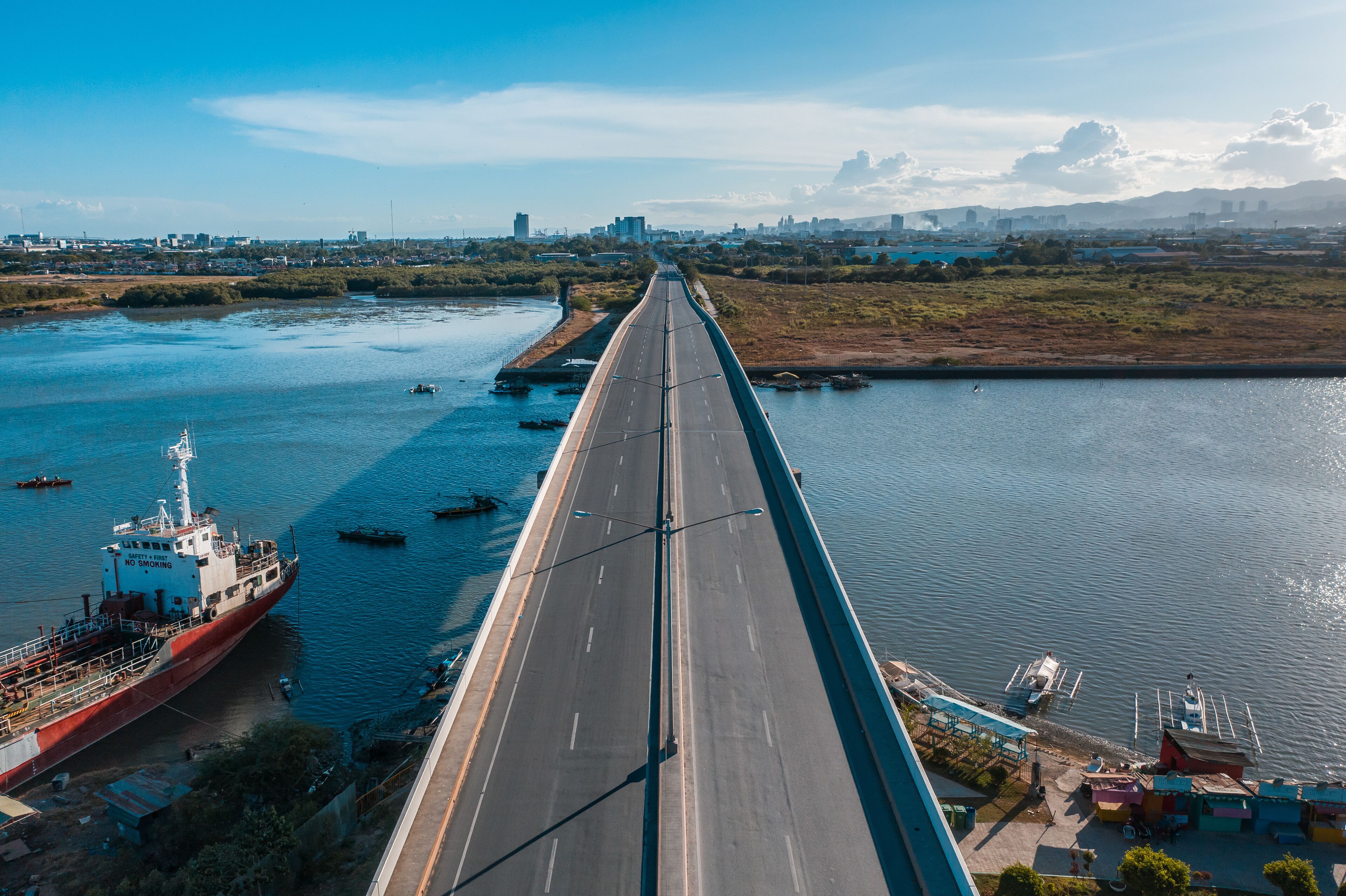 A long bridge over a river with boats and a cityscape in the background.