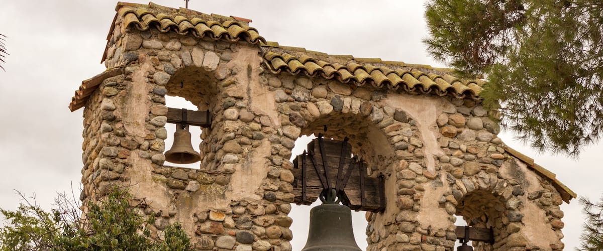 A stone bell tower with three bells, surrounded by trees.