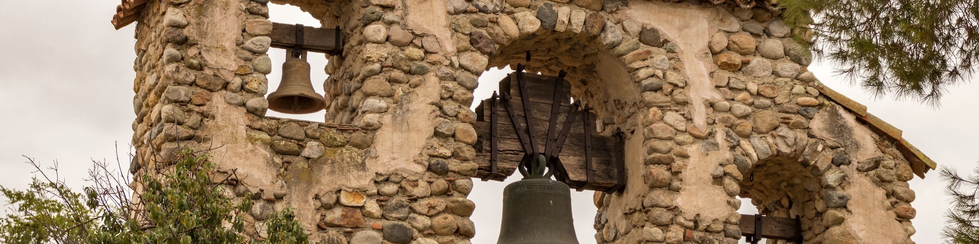 A stone bell tower with three bells, surrounded by trees.