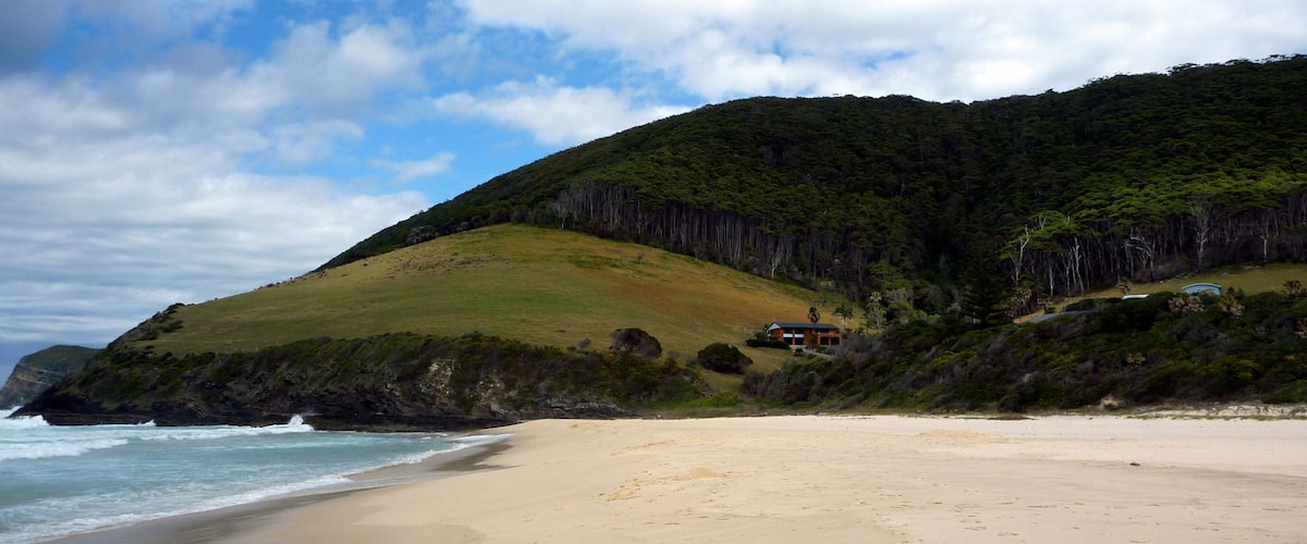 A beach with a house on a hill, ocean waves, and a forested hillside.