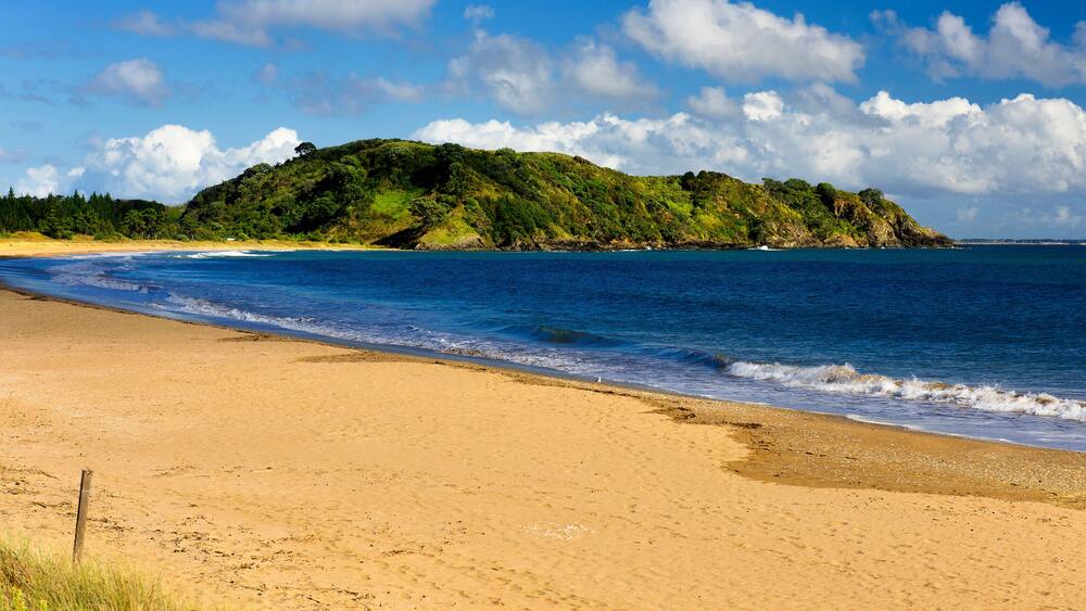 A sandy beach with gentle waves, a clear blue sky, and a green hill in the background.