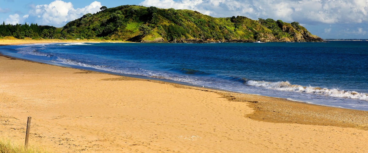 A sandy beach with gentle waves, a clear blue sky, and a green hill in the background.