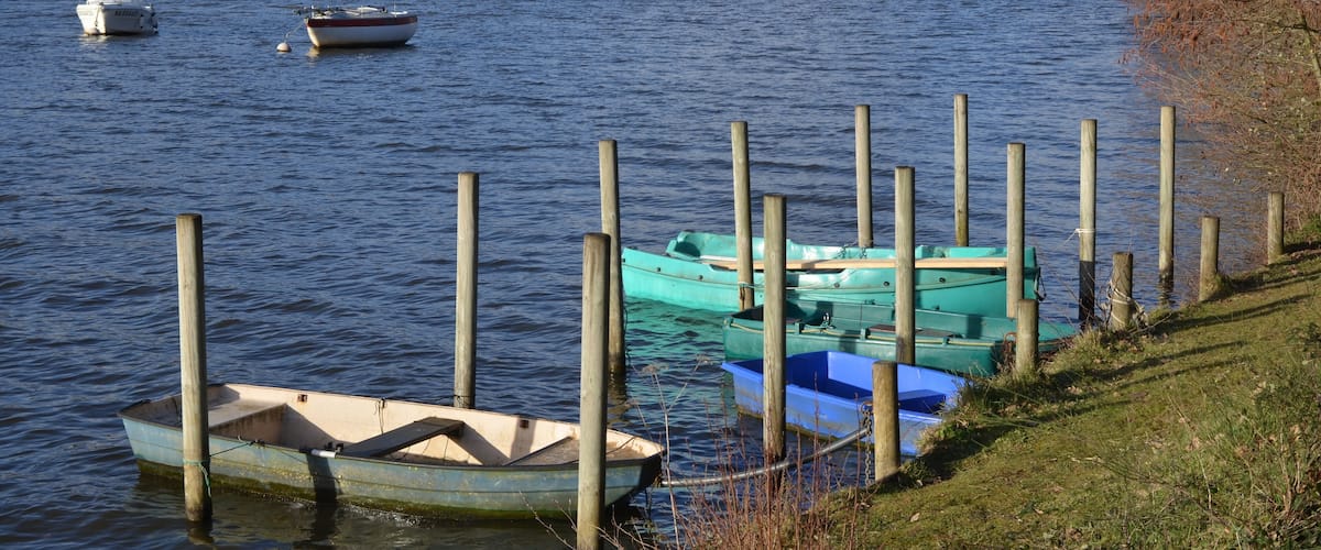 A calm river with boats docked at wooden piers, a bridge in the background, and a forested shoreline.