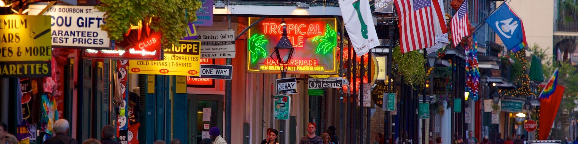 Uma rua movimentada com diversas lojas e bandeiras, incluindo um letreiro de neon para Tropical Island e várias bandeiras americanas.
