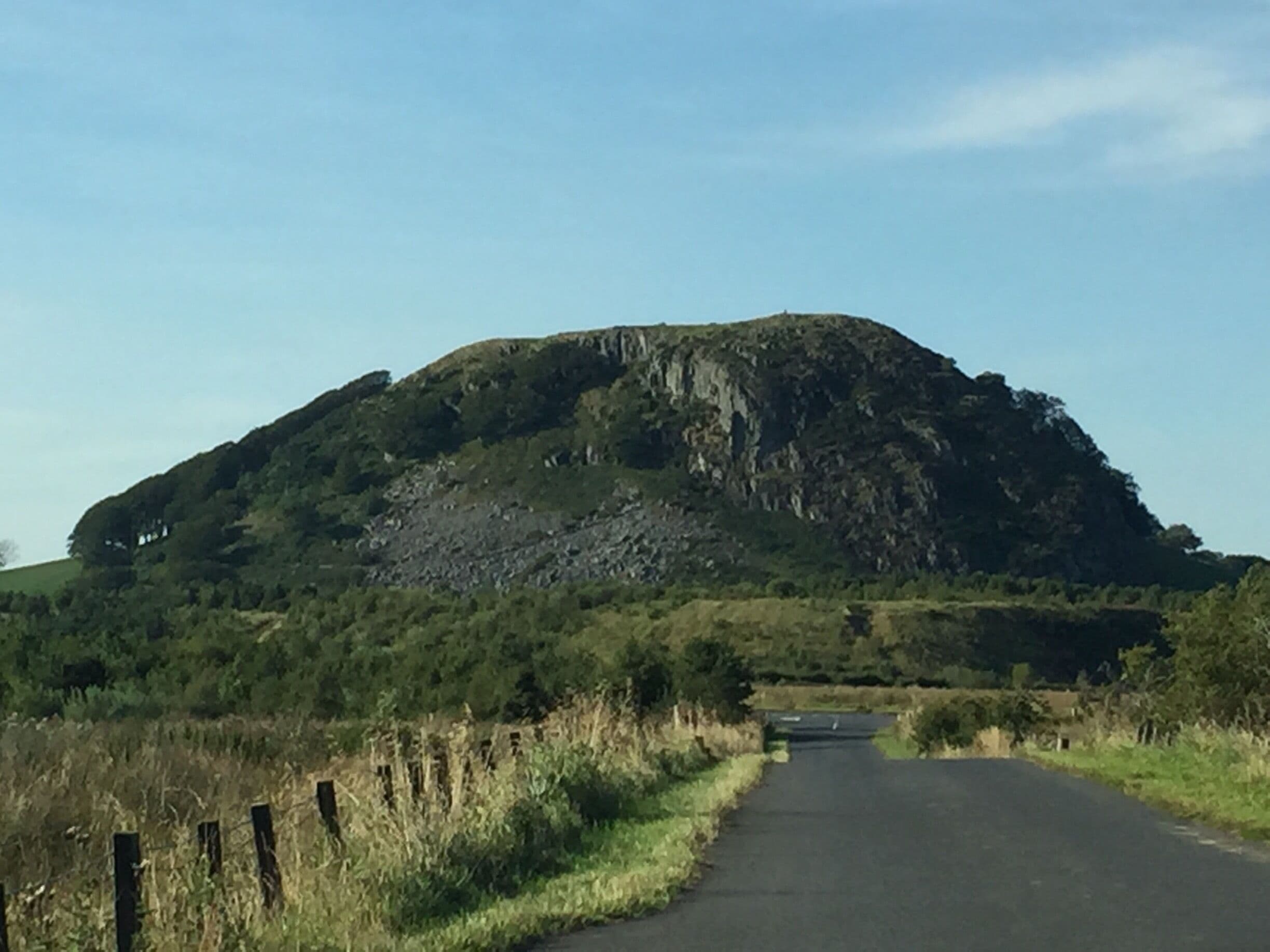 A rural road flanked by grassy fields and a large hill with a rocky outcrop.
