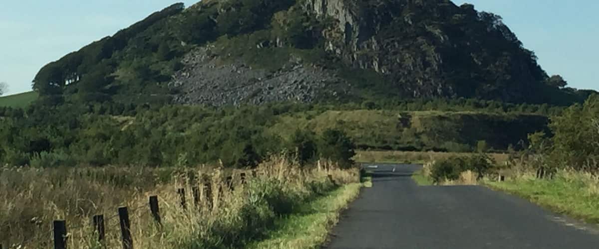 A rural road flanked by grassy fields and a large hill with a rocky outcrop.